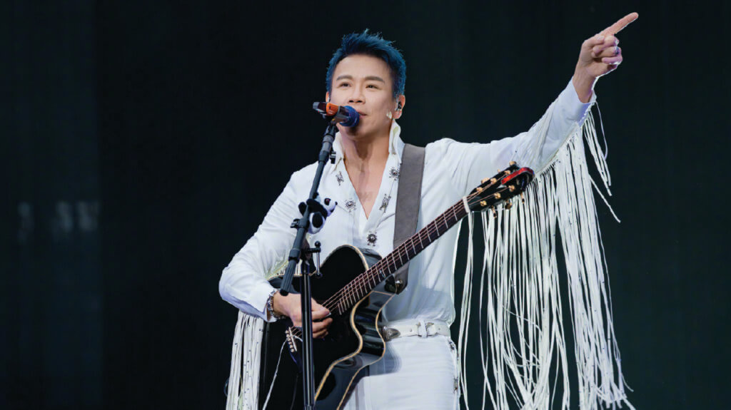 Singer David Tao, in a white outfit, holds a guitar and points, connecting with his audience during a performance.
