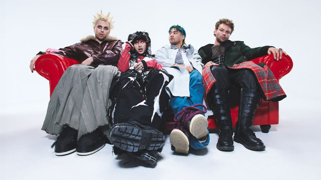 Members of 5 Seconds of Summer sitting together on a red couch, smiling and engaging with each other.