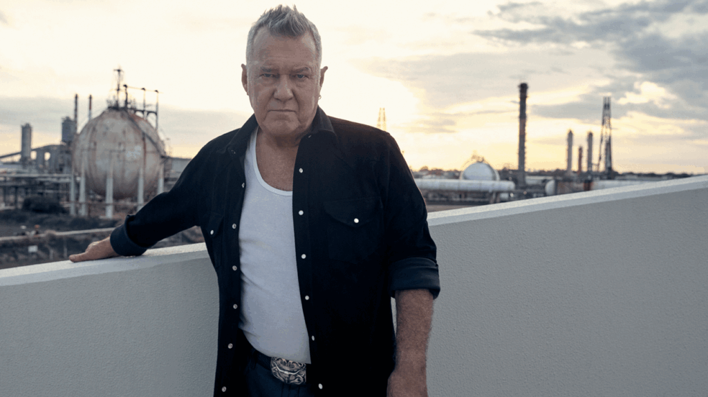 Singer Jimmy Barnes dressed in casual clothing posing against an industrial backdrop.