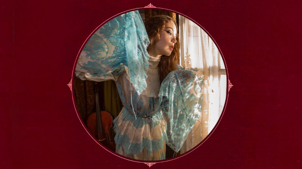 Laufey in a blue dress stands gracefully in front of a large window.