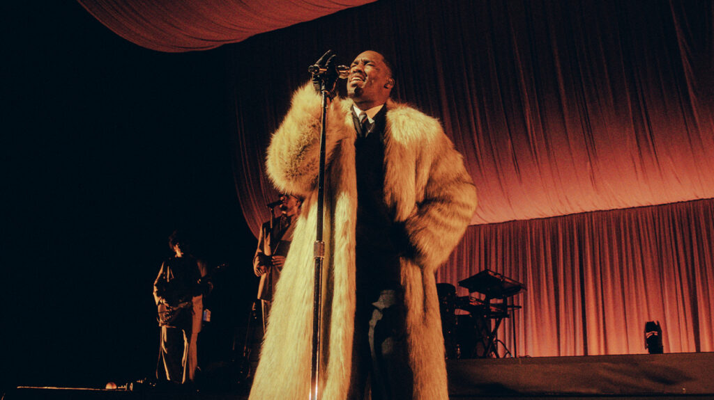 Singer GIVĒON dressed in a suit and fur coat, singing on stage.