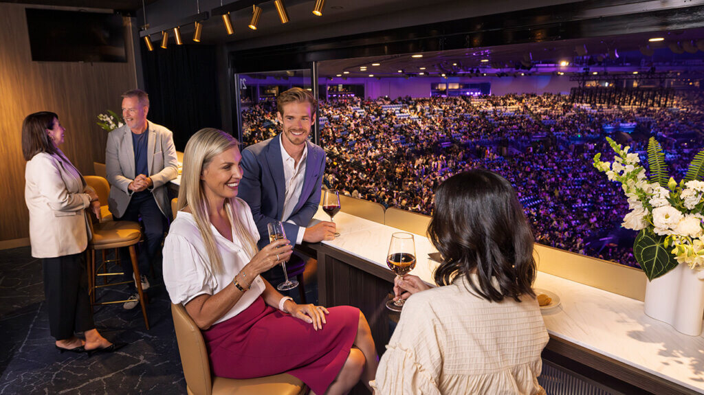 A group of people enjoying wine at a Rod Laver Arena suite, with a large crowd gathered in the arena.
