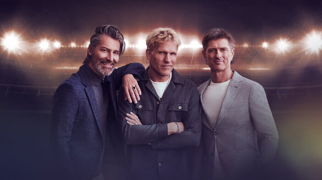 Three men from the band Michael Learns to Rock stand together in front of a large stadium.