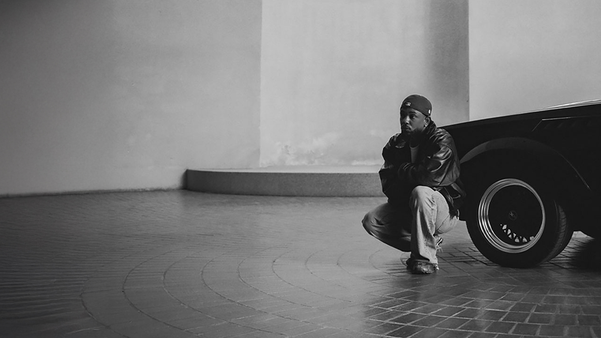 Kendrick Lamar kneels beside a car in a room.