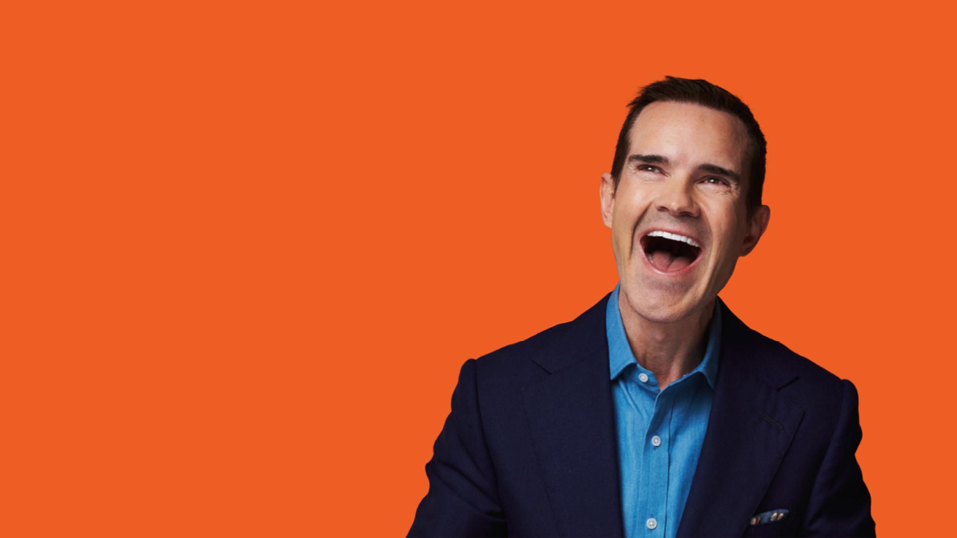 Comedian Jimmy Carr with mouth open laughing against an orange background.