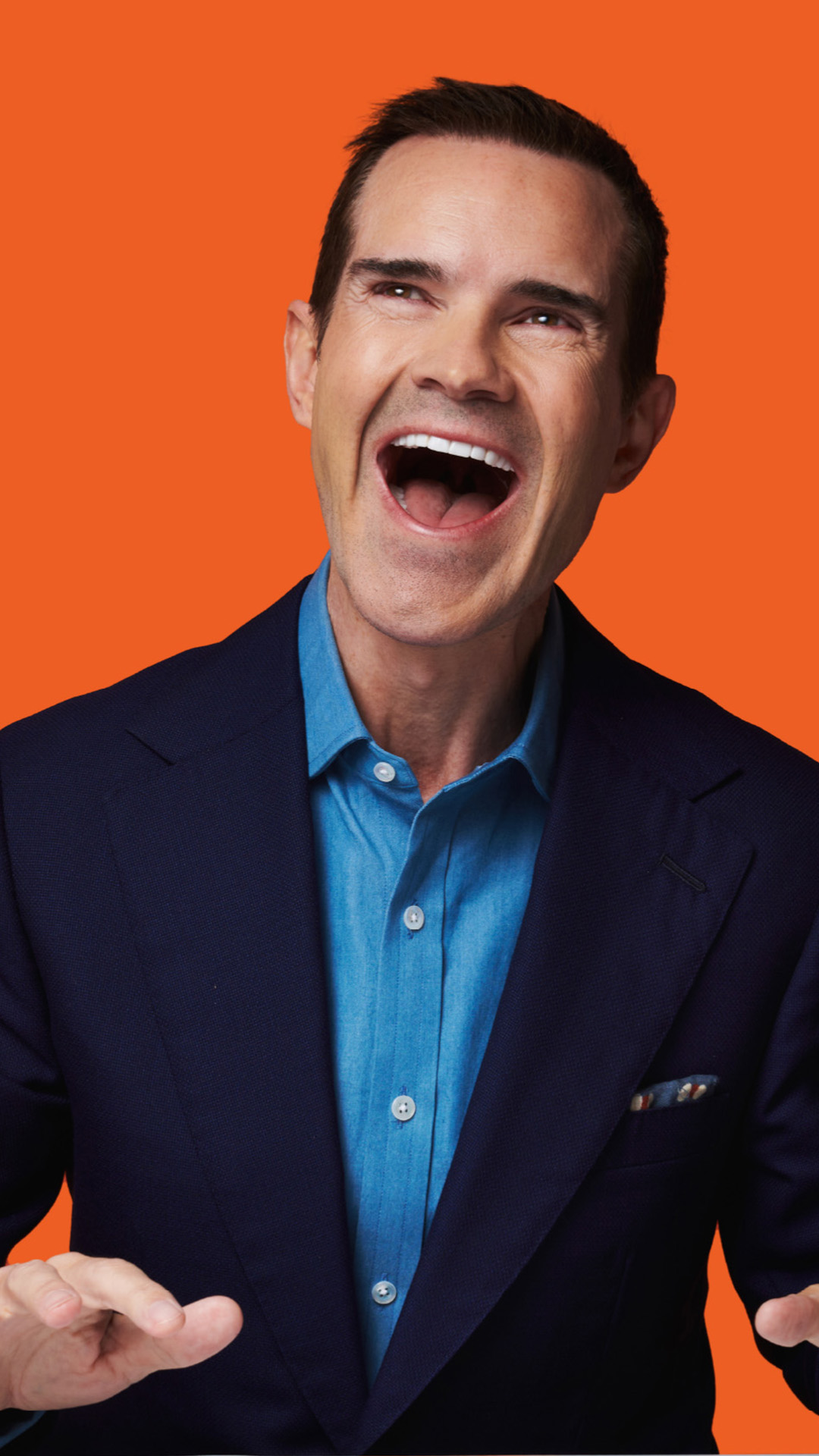 Comedian Jimmy Carr with mouth open laughing against an orange background.