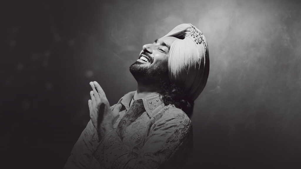 Singer Satinder Sartaaj with hands-clapped and a smiling pose against a grey background.