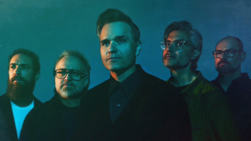 Death Cab For Cutie band members dressed in black and posing against a blue background.