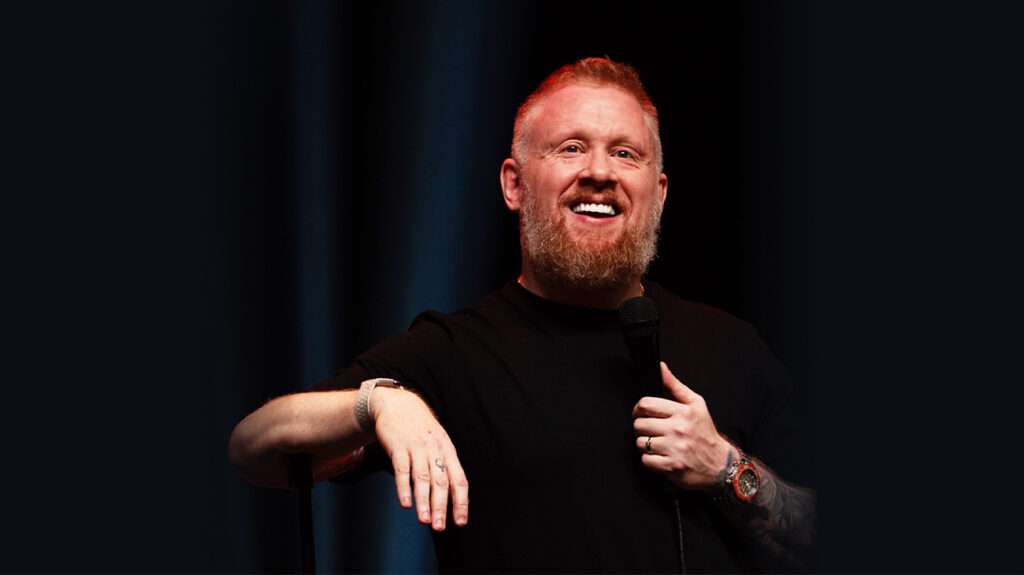 Paul Smith smiling brightly on stage with a microphone in hand.