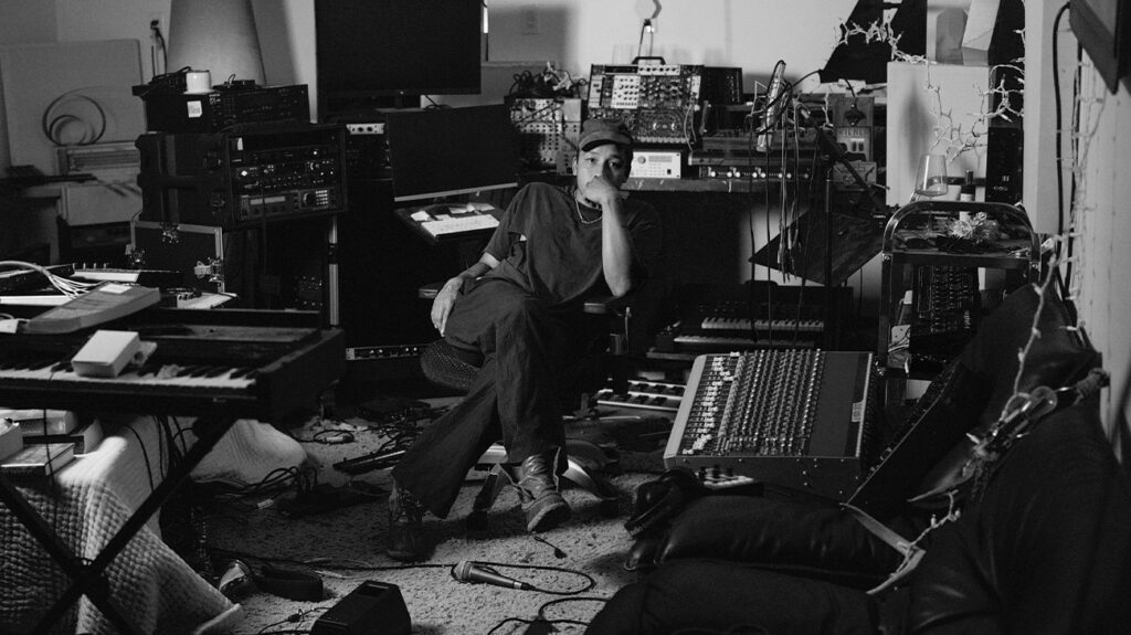 Dijon seated in a room filled with various electronic and musical equipment.