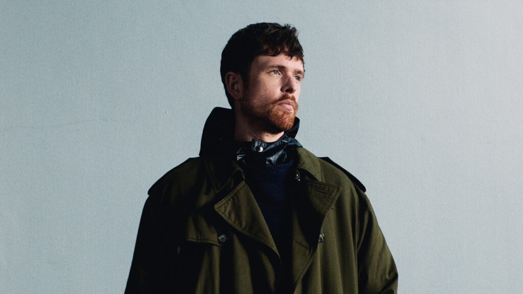 James Blake dressed in a green coat poses, highlighting his fashionable look against a plain background.