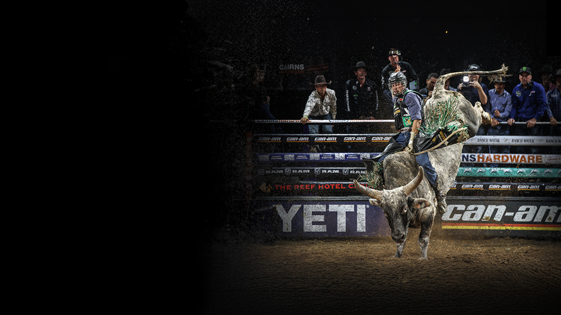 A man in a cowboy hat rides a bucking bull in a rodeo arena, surrounded by cheering spectators.