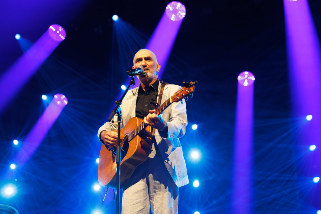 Paul Kelly performing on stage with a guitar during a concert.