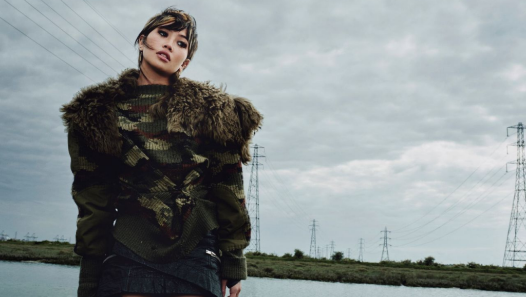 beabadoobee dressed in a camouflage jacket stands beside a body of water.