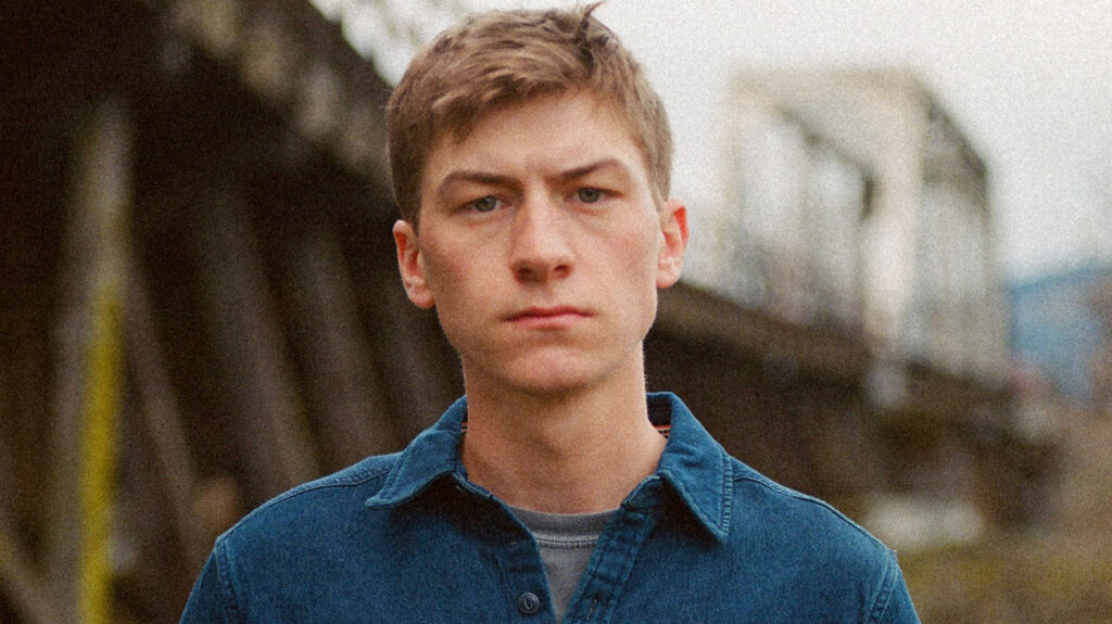 Sam Barber in a denim shirt posing seriously in an outdoor setting.
